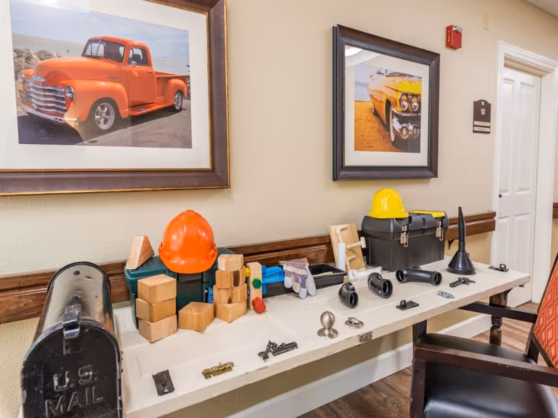 A hallway table displaying various tools and construction-related items including an orange hard hat, a yellow hard hat, wooden blocks, gloves, a toolbox, plumbing parts, and a black mailbox. Two framed pictures of vintage vehicles hang on the wall above the table.