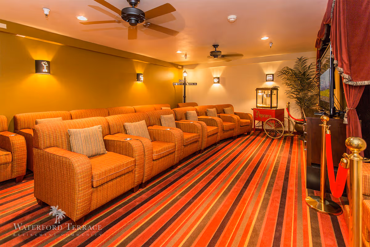 A cozy theater room with multiple orange upholstered armchairs arranged in rows facing a large screen. The room features a striped carpet in shades of red, orange, and brown, ceiling fans, wall sconces for lighting, a popcorn machine on a cart, and red velvet ropes with gold stanchions near the screen. There is a potted plant in the corner and the walls are painted in warm tones.