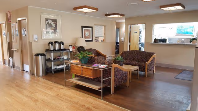 A nursing center common area with two patterned armchairs and a small wooden table between them. There is a wooden console table with plants and magazines on it. The room has light-colored walls, wooden flooring, and ceiling lights. To the left, there is a hallway with doors and a cart with coffee dispensers and supplies. On the right side, there is a reception window with a counter and a door nearby.