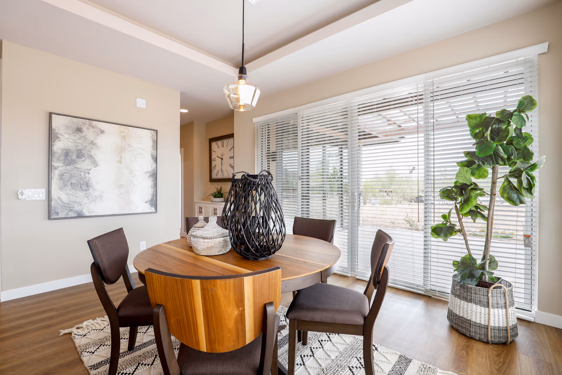 A bright dining area with a round wooden table surrounded by five dark cushioned chairs. On the table are decorative vases, including a large black woven vase and two smaller white ones. The room features large windows with white blinds, allowing natural light to fill the space. A tall green plant in a woven basket is placed near the window. The walls are light beige, adorned with a large abstract painting and a clock visible in the background. The floor is wooden with a patterned rug under the table.