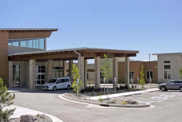 Exterior front entrance of a senior living building with a covered porte-cochere, landscaping, and parked vehicles.