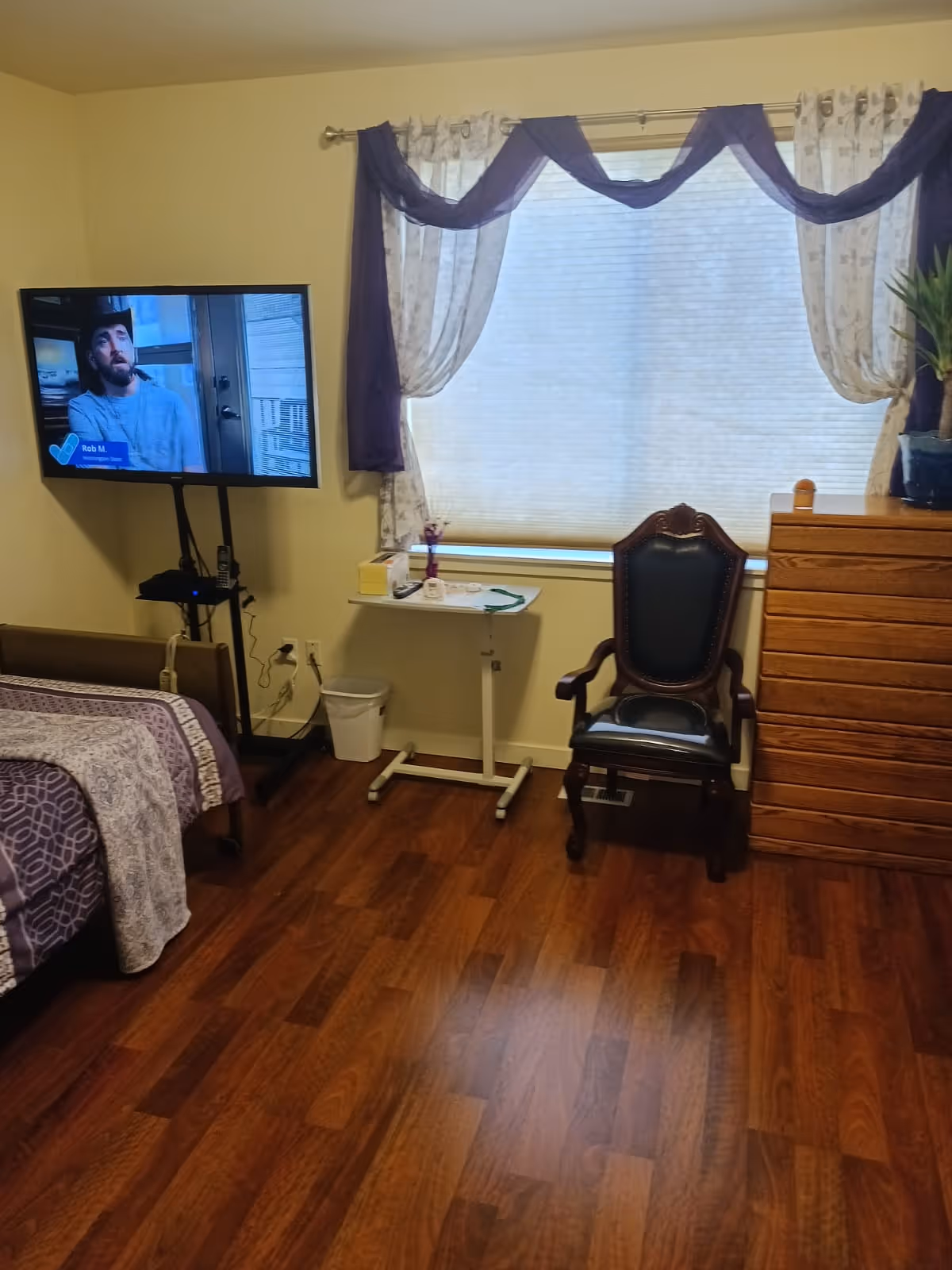 A senior bedroom with a bed, wall-mounted TV, wooden dresser, chair, small rolling table and a curtained window.