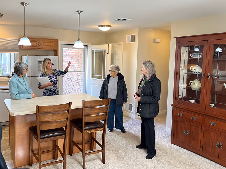 Four women standing and talking in a kitchen area with a large island countertop, two wooden chairs, pendant lights, a refrigerator, and a wooden china cabinet displaying dishes.