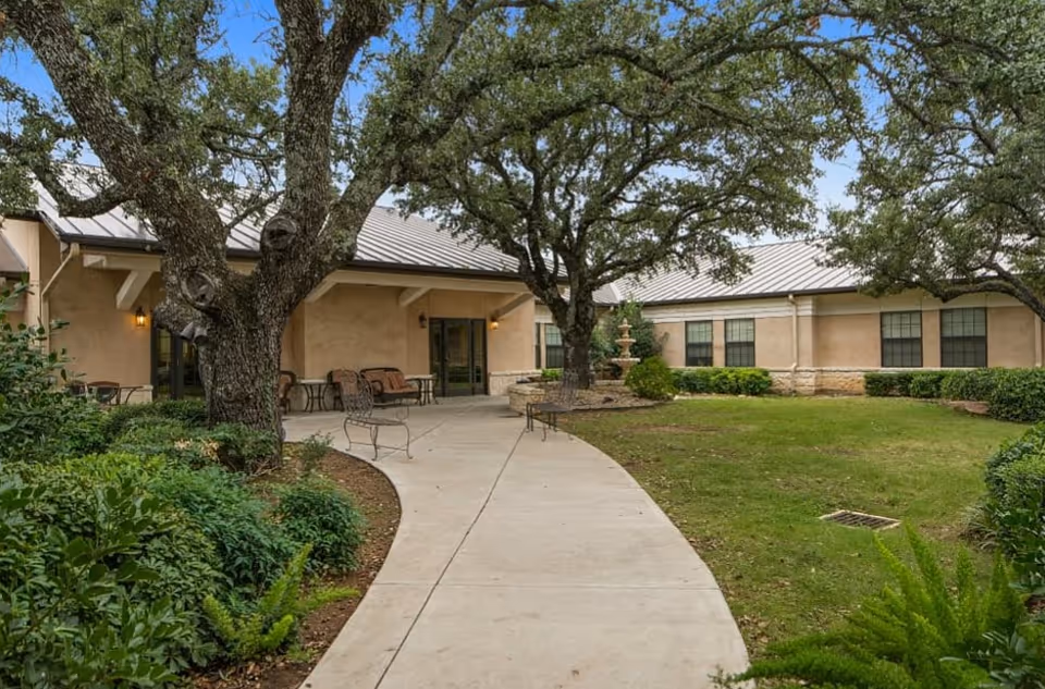 Outdoor courtyard area at Arbor House Assisted Living and Memory Care - Llano featuring a curved concrete walkway, mature trees, green shrubs, and a beige building with multiple windows and a metal roof. There are benches and small tables along the walkway near the building entrance.
