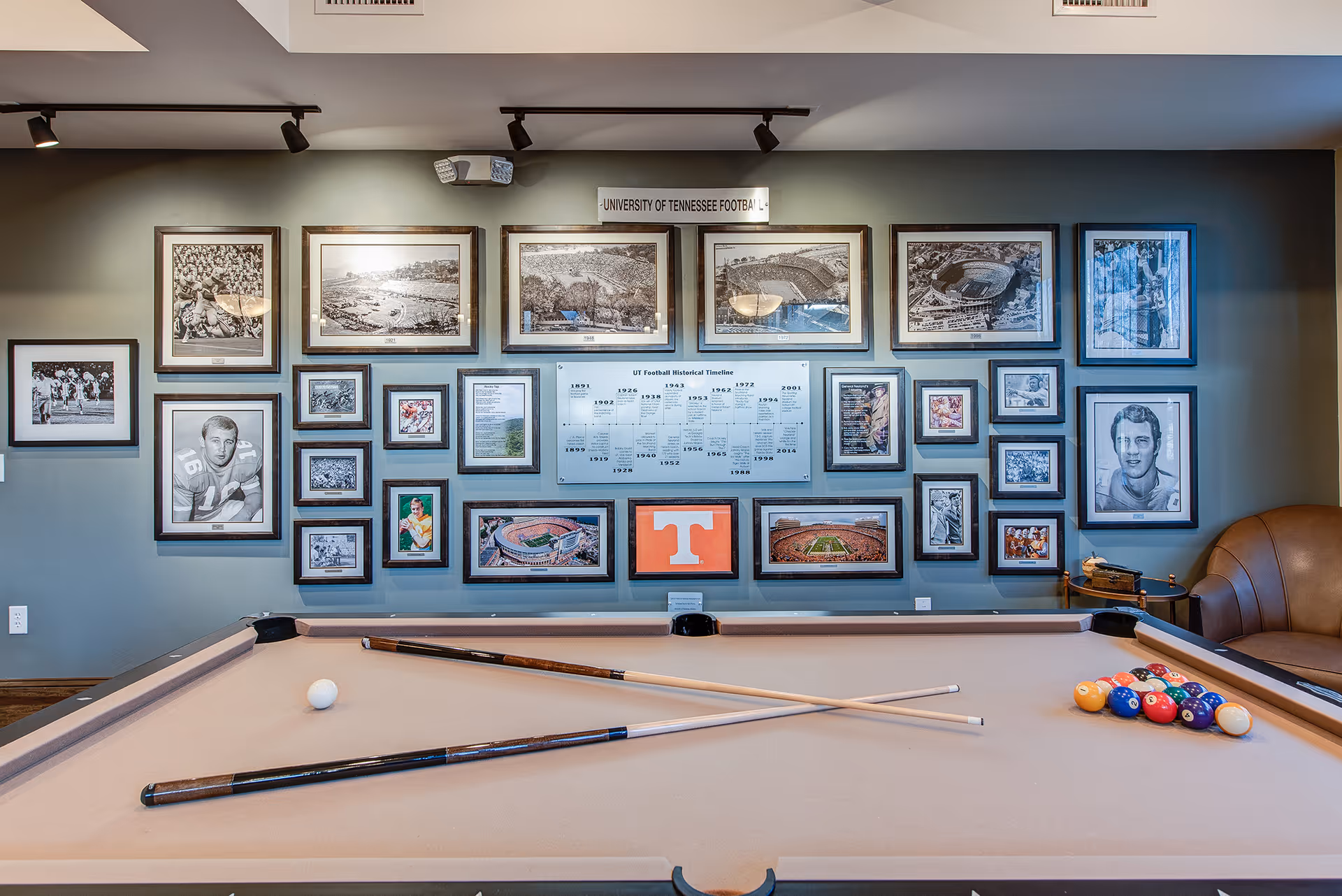 A billiards table with cues and racked balls in front of a wall of framed sports photos and memorabilia, including a University of Tennessee 'T' logo.