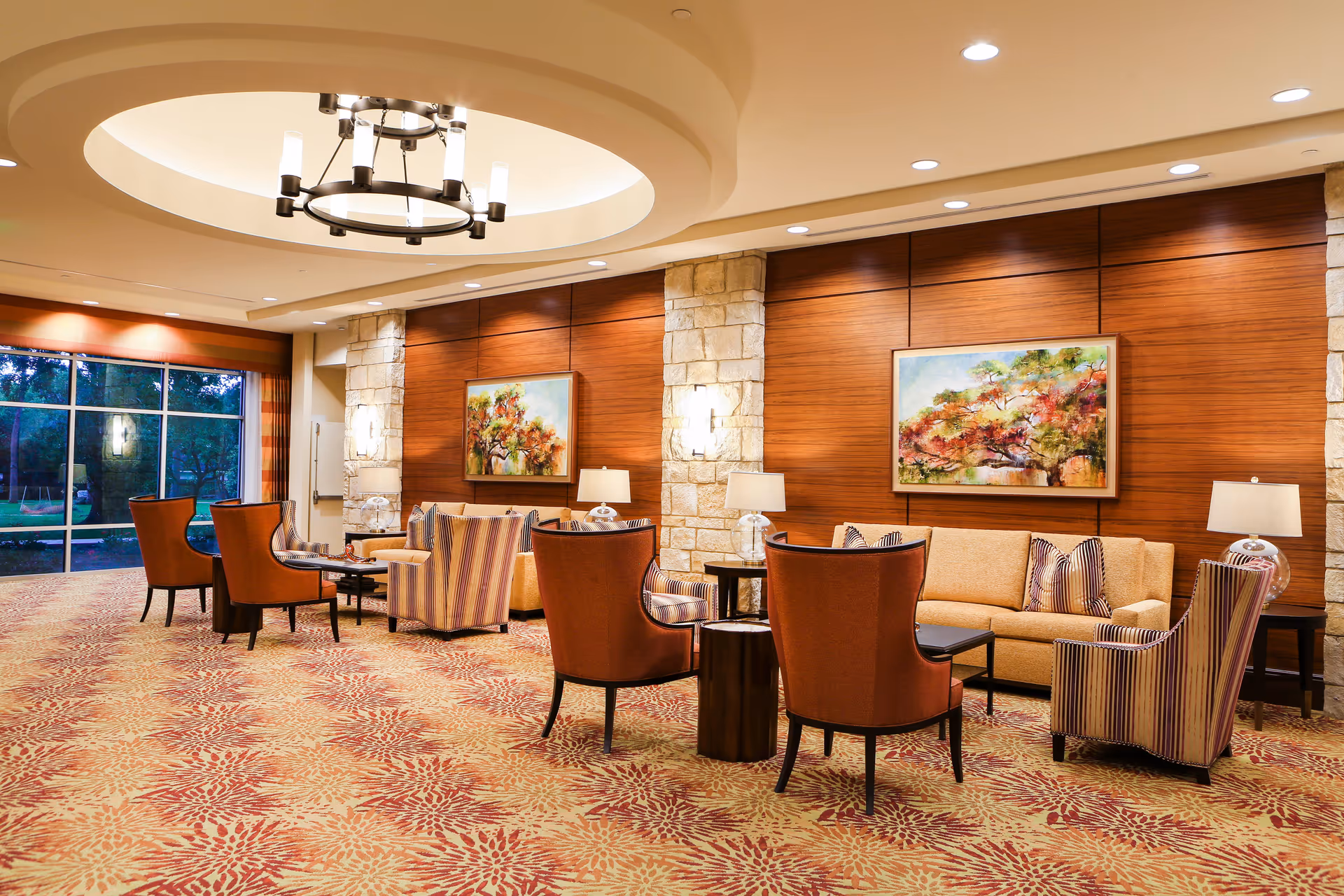 A spacious and well-lit lounge area with comfortable seating including armchairs and sofas arranged around small tables. The room features a patterned carpet, wooden paneled walls with stone accents, two colorful paintings, table lamps, and a large circular ceiling light fixture. Large windows on one side provide a view of greenery outside.