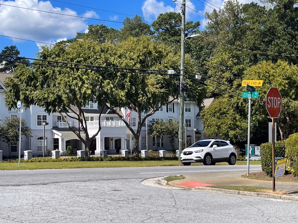 Street view of a white multi-story senior living building with large trees, a parked white SUV, and a stop sign at an intersection.
