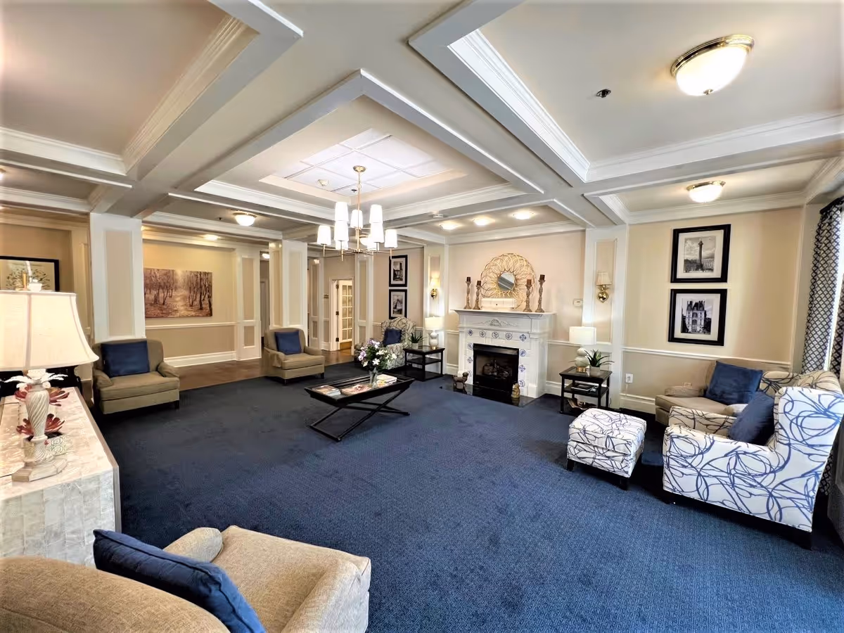 A spacious and well-lit living room with a blue carpet, beige walls, and white coffered ceiling. The room features several armchairs with blue and beige cushions, a coffee table with flowers and magazines, a white fireplace adorned with decorative items, framed artwork on the walls, and multiple lamps providing warm lighting.