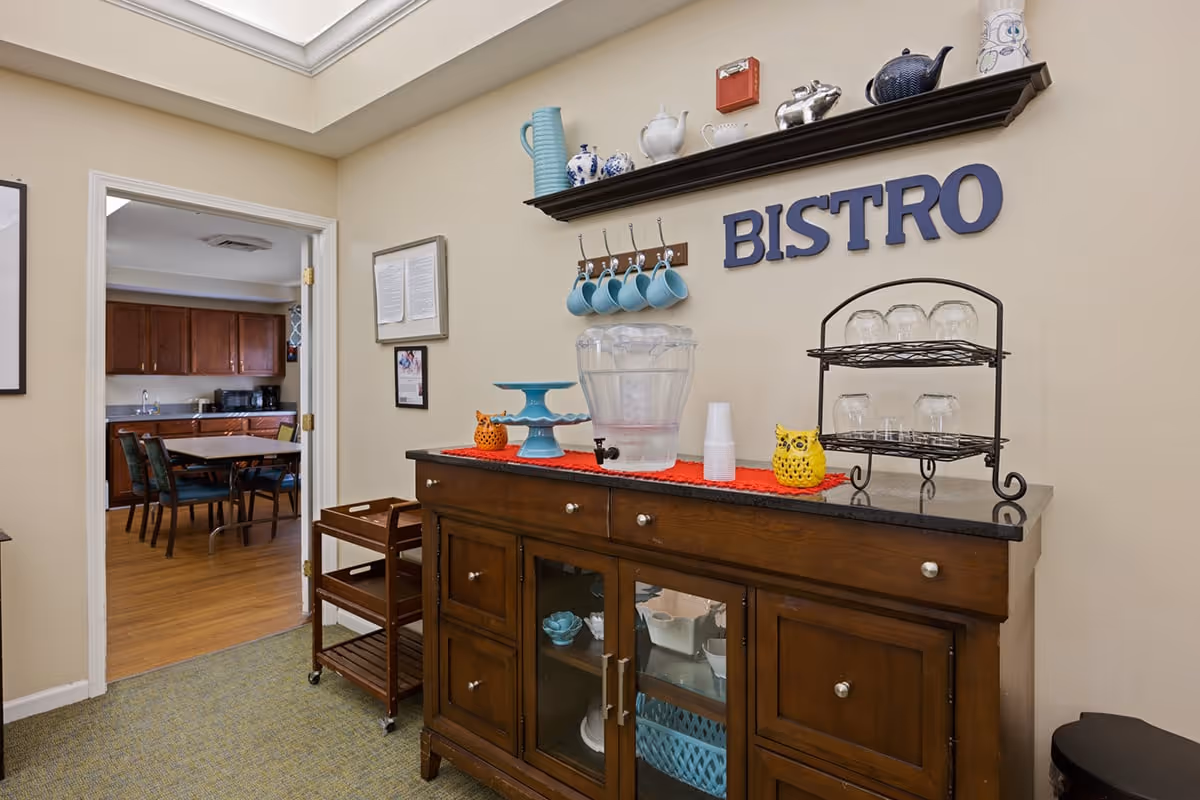 Interior view of a bistro area in a senior living facility with a wooden cabinet holding a water dispenser, cups, and decorative items. Above the cabinet, there is a shelf with teapots and mugs hanging on hooks. The word 'BISTRO' is displayed on the wall. Through an open doorway, a kitchen or dining area with a table and chairs is visible.