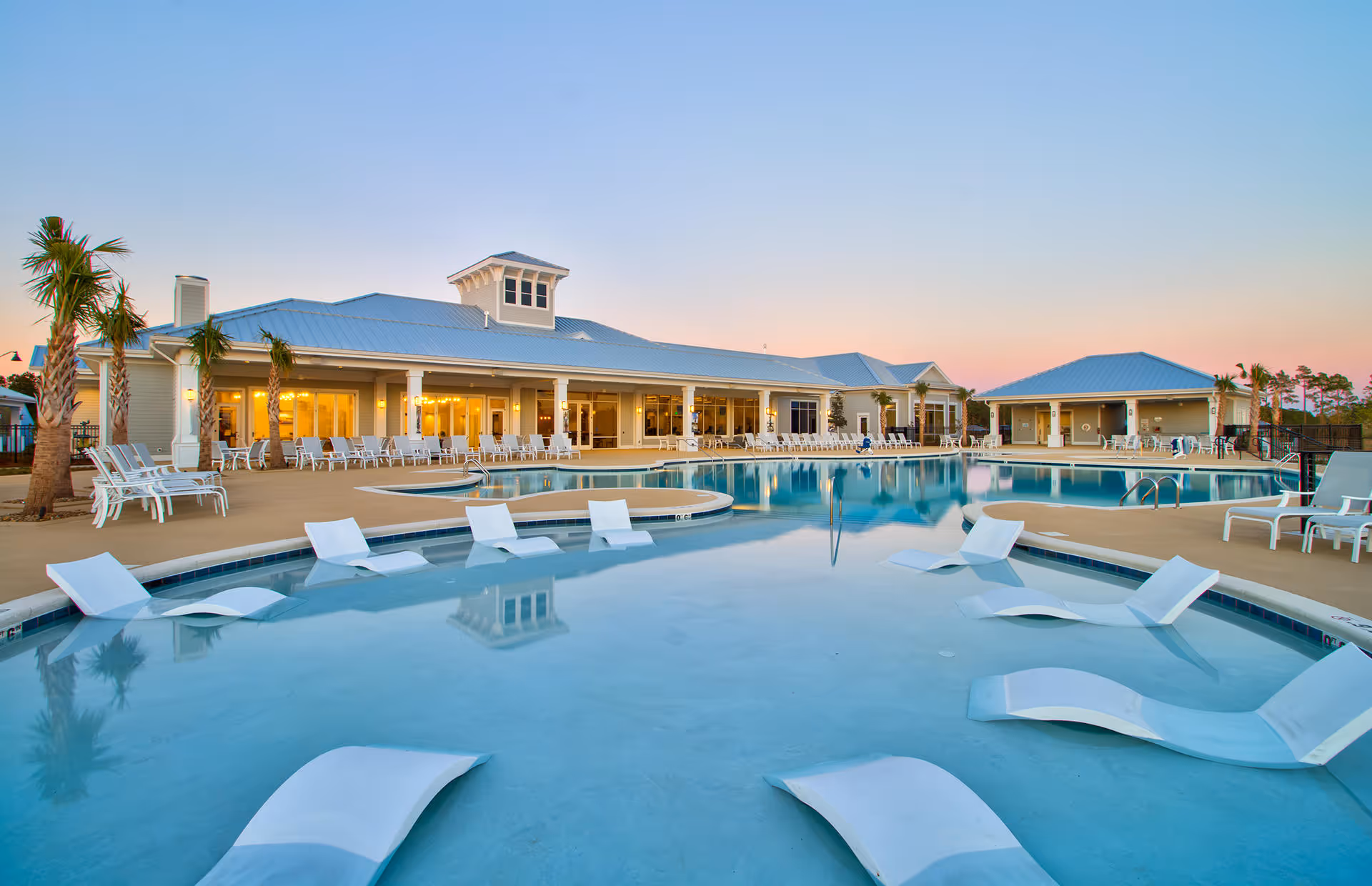 Outdoor swimming pool area at Del Webb Wilmington 55+ Retirement Community during sunset, featuring lounge chairs partially submerged in the pool, palm trees, and a large building with a covered patio and many chairs around the pool deck.