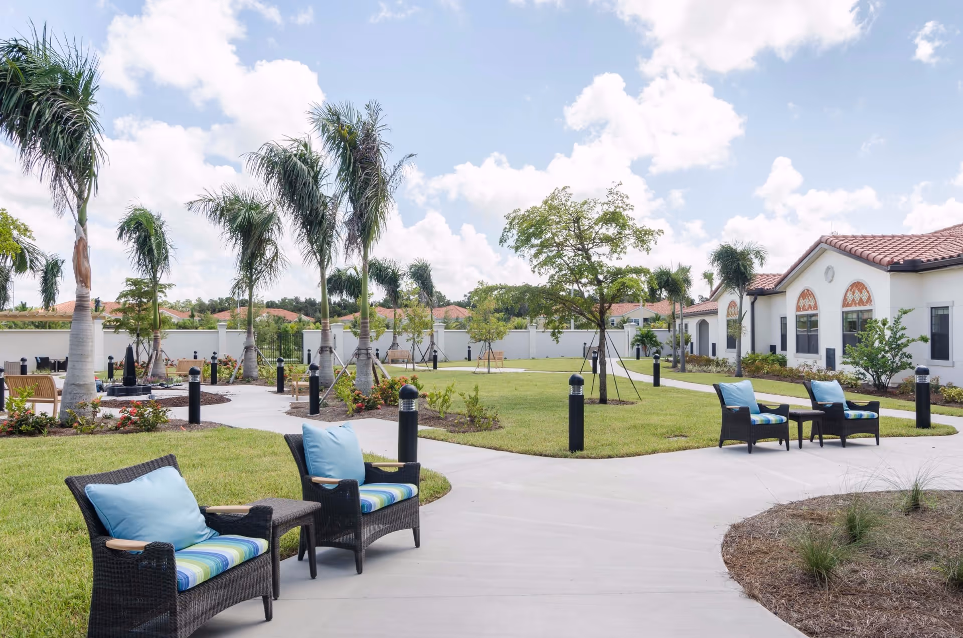 Sunny outdoor courtyard with wicker seating, palm trees, paved walkways and the facility building in the background.