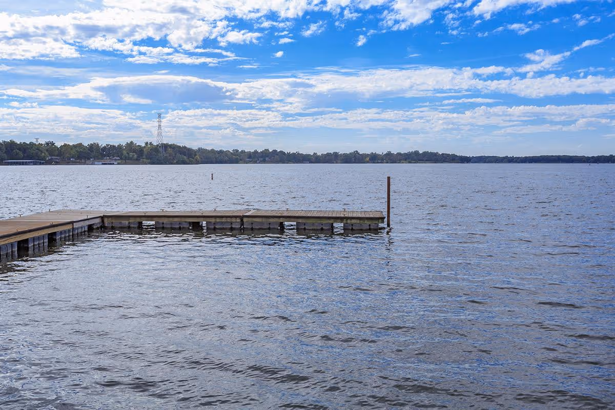 A wooden dock extending into a large body of water under a partly cloudy blue sky with a tree-lined shore in the distance.