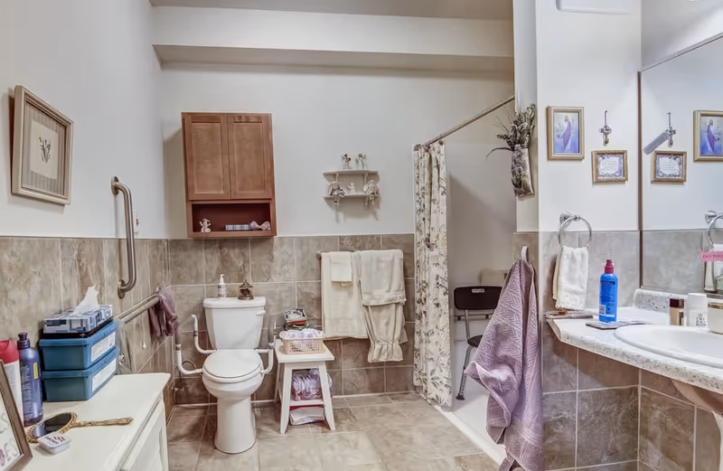A senior-friendly bathroom with a toilet equipped with support rails, a shower area with a curtain and a shower chair, a sink with a large mirror above it, and various towels and toiletries neatly arranged. The walls are tiled halfway up, and there is a wooden cabinet and small shelves for storage.
