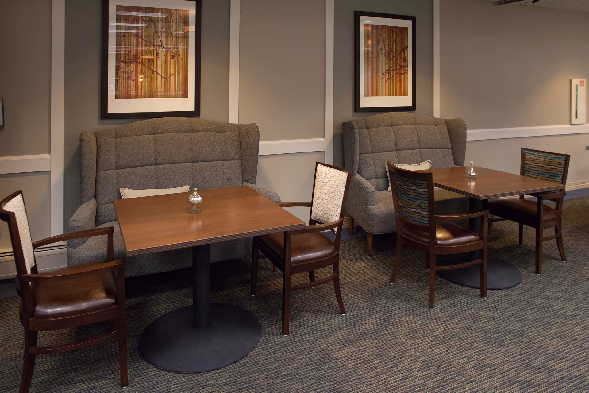 Two wooden tables each accompanied by two wooden chairs and a gray cushioned bench against a beige wall with framed artwork above. The setting appears to be a quiet seating or dining area with carpeted flooring.