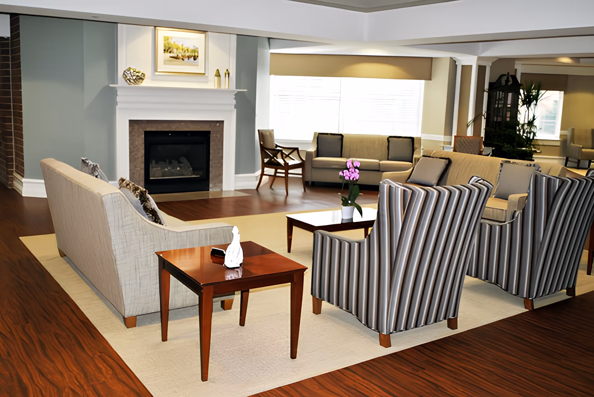 A spacious living room area with hardwood floors, a fireplace, and multiple seating options including beige sofas and striped armchairs arranged around a coffee table with a potted orchid. Large windows allow natural light to fill the room.