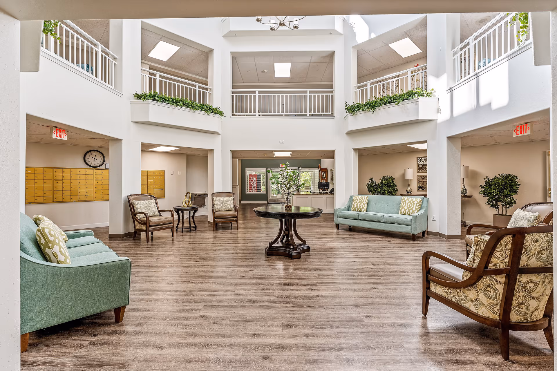 Spacious and bright common area in a senior living facility with light wood flooring, two light blue sofas with patterned pillows, several wooden armchairs with patterned cushions, a round wooden table with a flower arrangement in the center, and a second-floor balcony with white railings and green plants. There are exit signs and a wall clock visible, along with a set of mailboxes on the left wall.