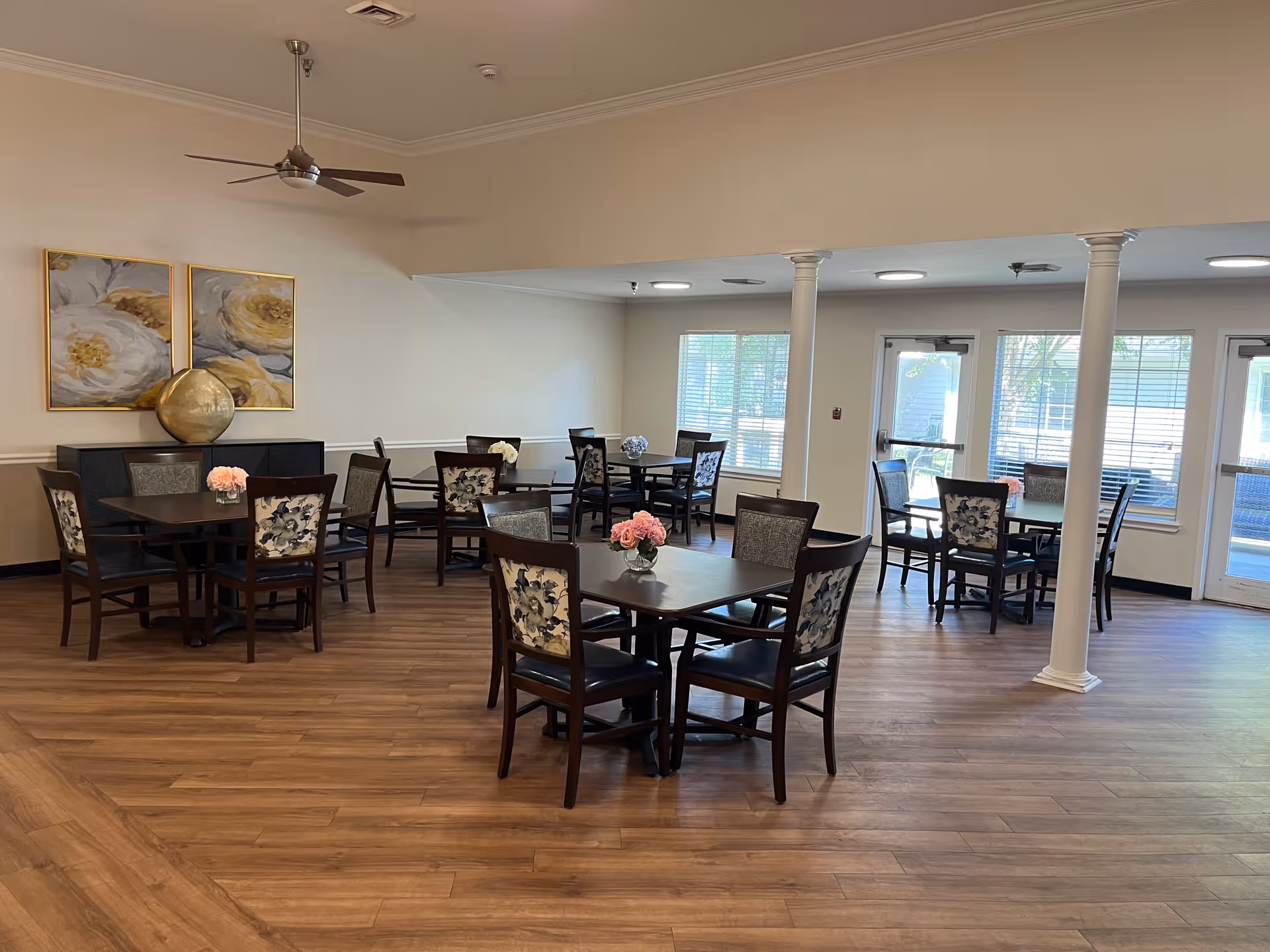 A well-lit dining room with several dark wooden tables and chairs arranged neatly. Each table has a small floral centerpiece. The room features wood flooring, white walls, two large floral paintings, a ceiling fan, and large windows letting in natural light. There are also white columns and glass doors leading outside.
