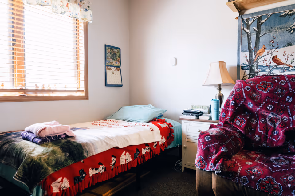 Sunlit small bedroom with a single bed, nightstand with a lamp and personal items, and a patterned armchair.