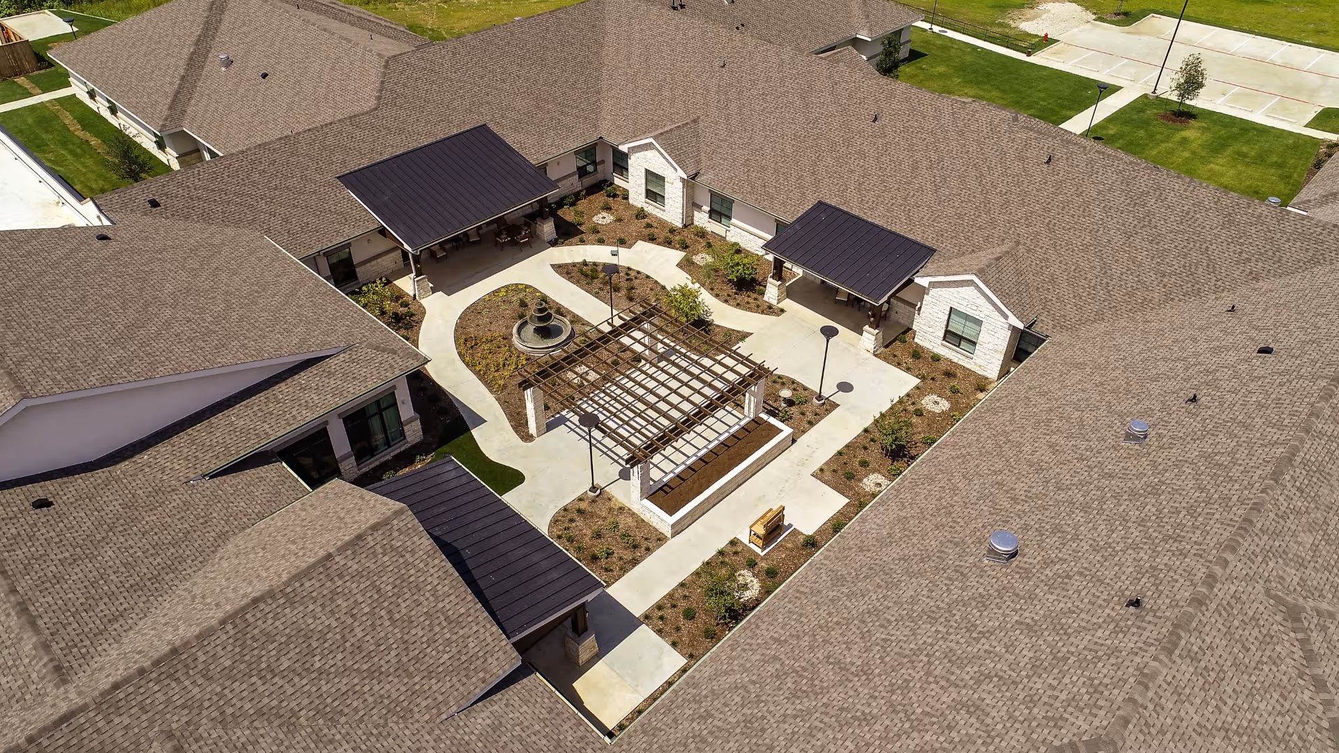Aerial view of an outdoor courtyard area within a senior living facility. The courtyard features a central pergola structure, a circular fountain, paved walkways, landscaped garden beds, and covered seating areas attached to the surrounding building. The building has a brown shingled roof and light-colored exterior walls.