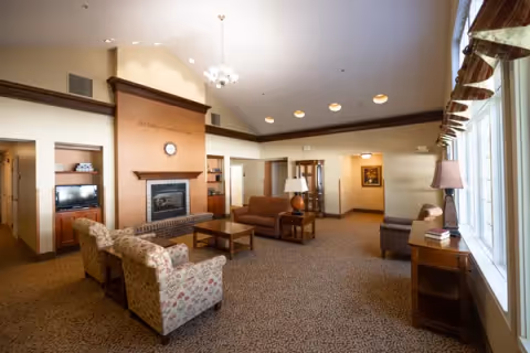 A spacious senior living common area with a fireplace, several armchairs and sofas arranged around coffee tables, a TV in a wooden cabinet, large windows with curtains letting in natural light, and warm beige walls with decorative molding.