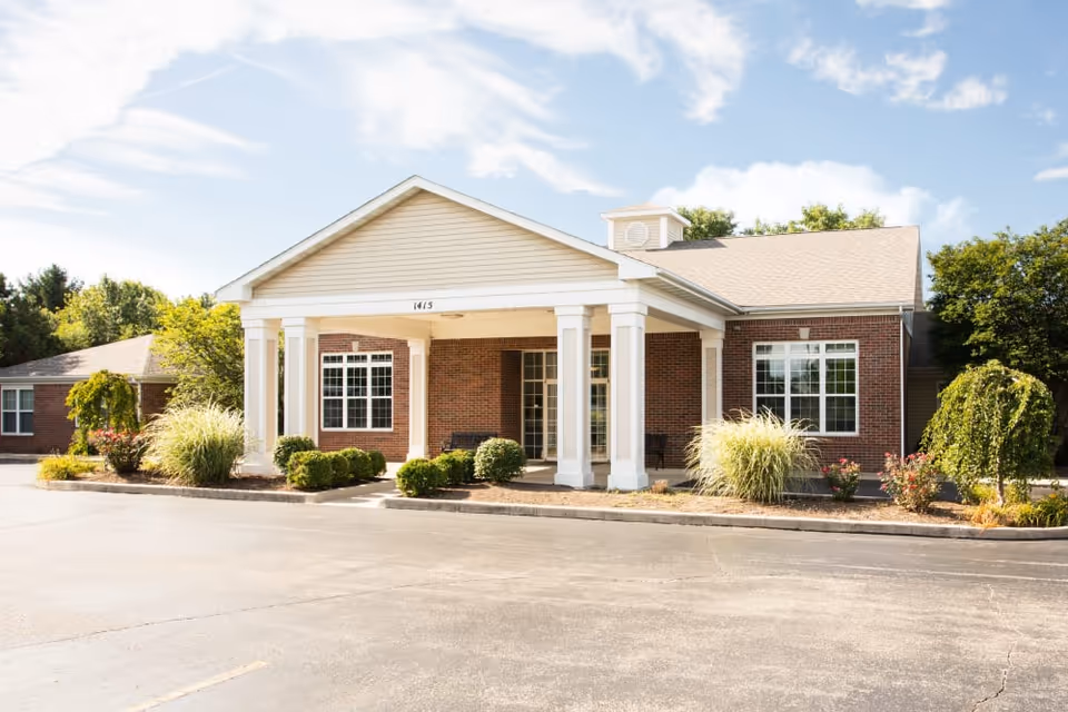 Exterior front view of a single-story brick building with white columns supporting a covered entrance. The building is surrounded by landscaped bushes and plants, with a clear blue sky overhead.