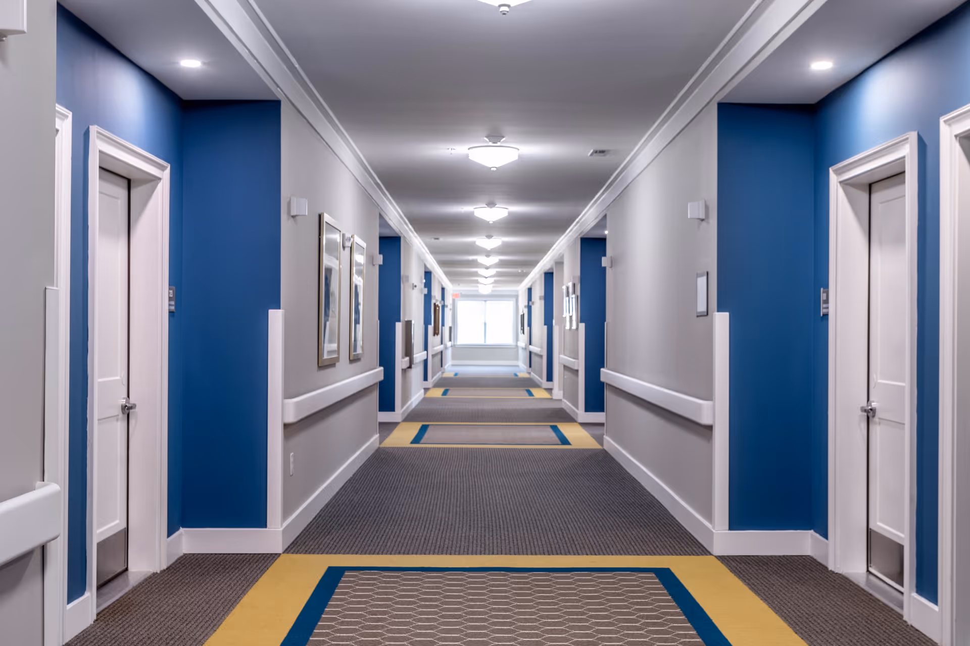 Long carpeted interior hallway with blue accent walls, white doors, handrails, and ceiling lights.