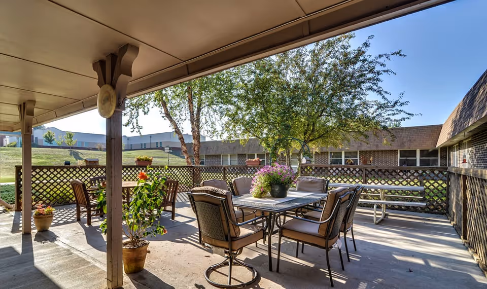 Covered outdoor patio with a dining table and chairs, potted plants, and lattice railing overlooking a grassy courtyard and adjacent buildings.
