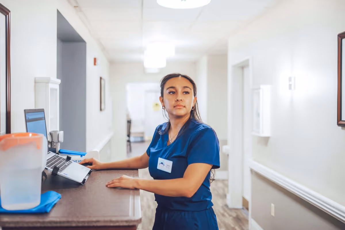 A woman in blue scrubs stands at a reception desk in a well-lit hallway of an assisted living facility, looking to her left. The desk has a laptop and a water pitcher on it, and the hallway has white walls with framed pictures and ceiling lights.