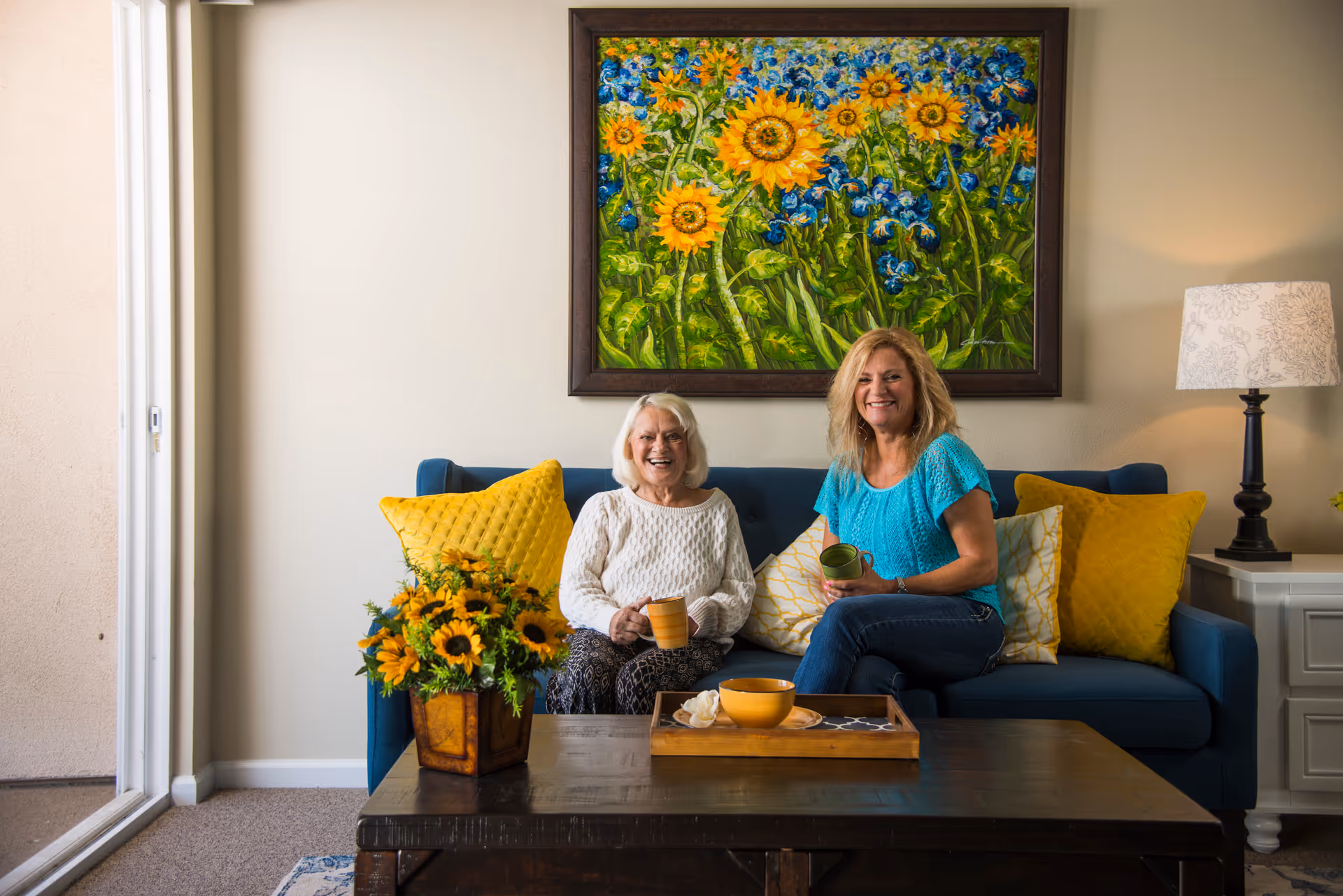 Two women sitting on a blue couch in a living room, smiling and holding mugs. The couch has yellow and white pillows, and there is a wooden coffee table in front of them with a tray and a bowl. A colorful painting of sunflowers and blue flowers hangs on the wall behind them, and a table lamp is on a side table to the right.