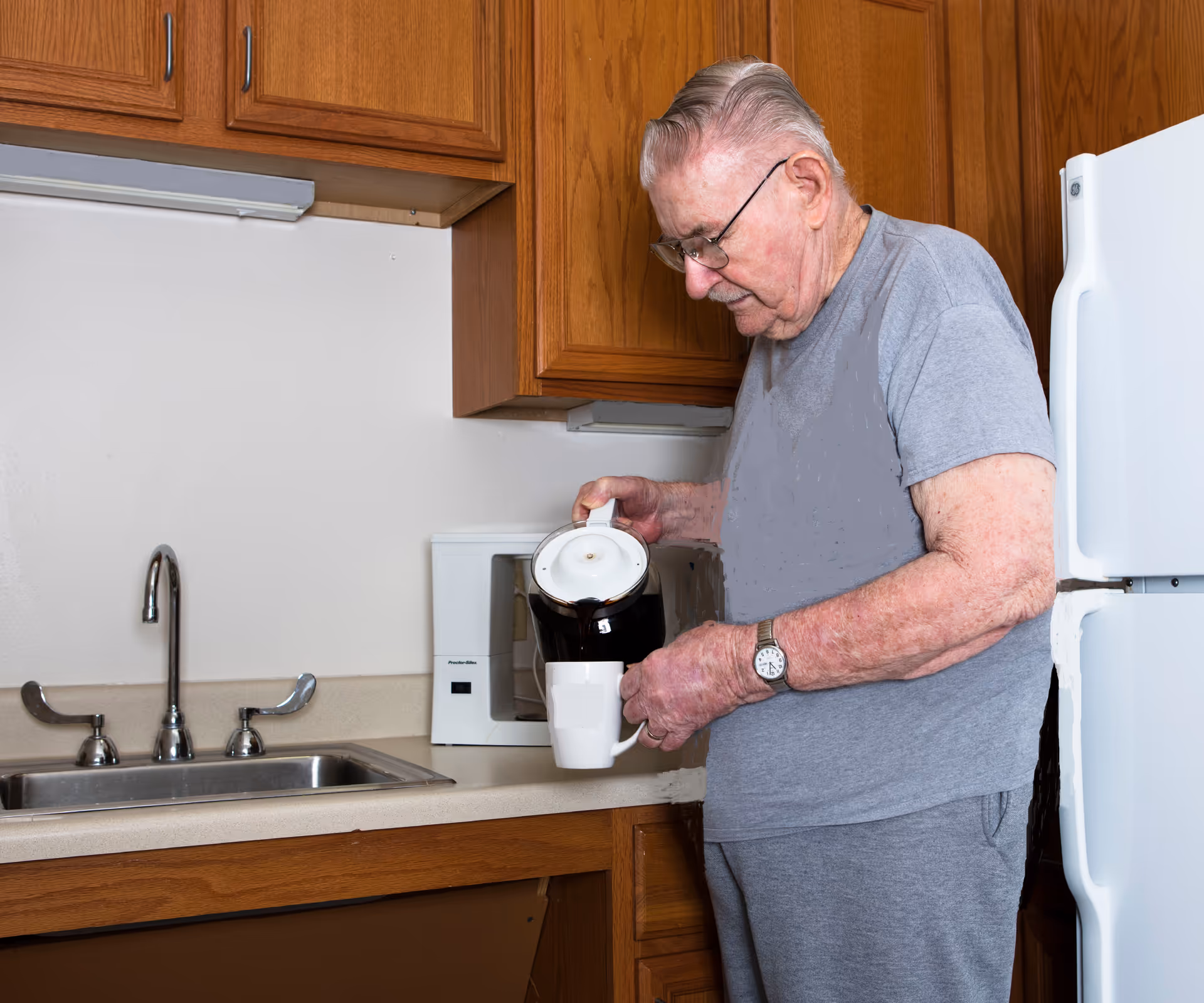An elderly man wearing glasses and a gray t-shirt is pouring coffee from a black coffee pot into a white mug in a kitchen with wooden cabinets, a white microwave, a white refrigerator, and a stainless steel sink.