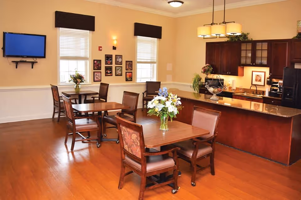 A warm and inviting dining area in a senior living facility featuring wooden tables and chairs with floral upholstery. Each table has a vase with fresh flowers. The room has hardwood floors, beige walls, two windows with blinds and valances, a wall-mounted TV, and a kitchen area with dark wood cabinets and a granite countertop. The kitchen area is decorated with plants and framed pictures.
