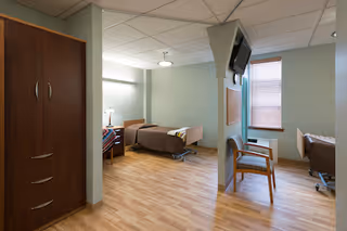 A spacious senior care facility room with two hospital-style beds separated by a partial wall. Each bed has a bedside table and lamp. There is a wooden wardrobe on the left side and a chair near the window with blinds. The room has light-colored walls and wood flooring, with a ceiling-mounted light fixture and a wall-mounted television.