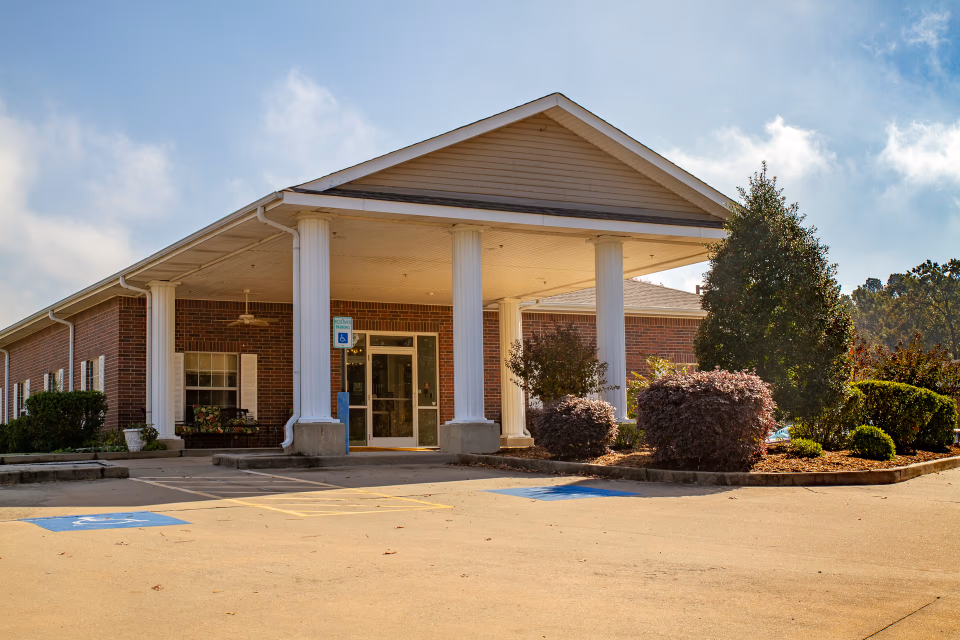 Exterior view of The Blossoms at White Hall Rehab & Nursing Center showing the front entrance with large white columns, a covered drop-off area, brick walls, windows with white shutters, and landscaped bushes and trees. The parking lot in front includes marked handicapped parking spaces.
