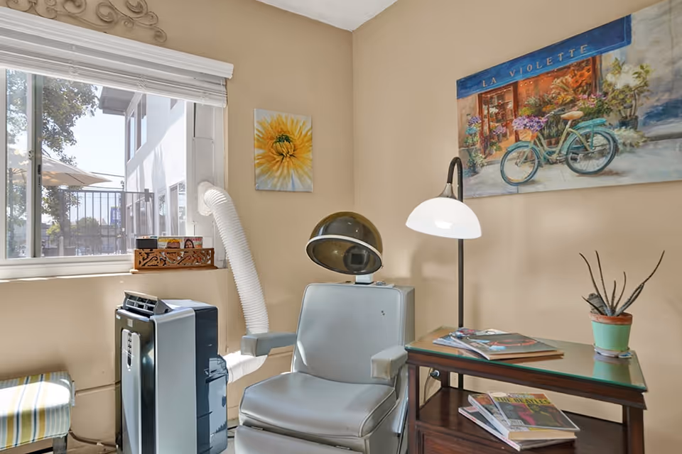 A cozy interior corner of a senior living facility room with a vintage hair dryer chair, a small table with magazines and a potted plant, a floor lamp, and two paintings on the beige walls. A window shows an outdoor view with trees and a building.