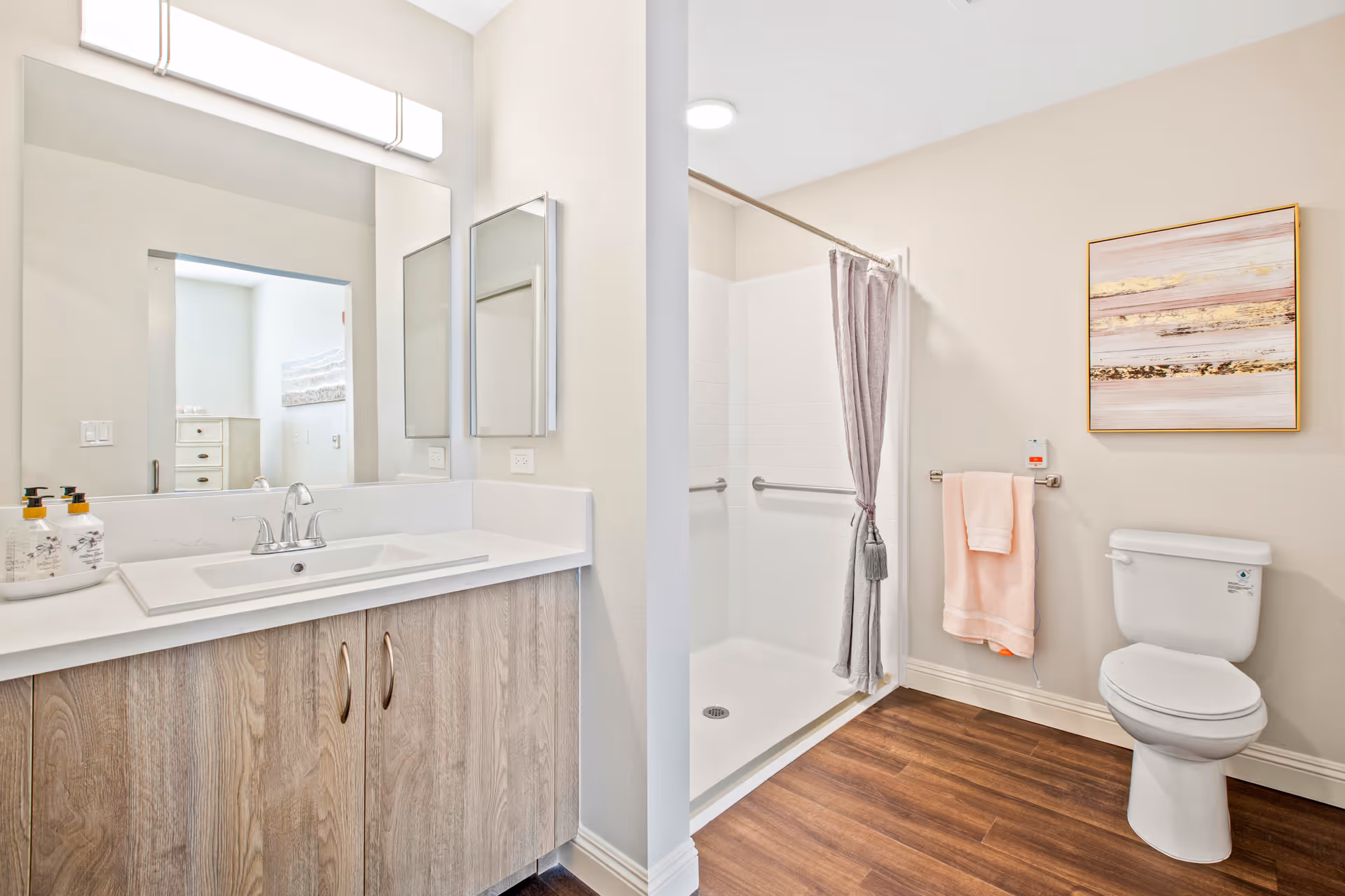 Bright modern bathroom with a sink vanity and large mirror, a walk-in shower, and a toilet with towel and wall art.