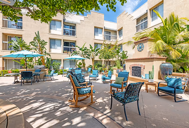 Sunlit courtyard with patio chairs, tables, umbrellas, and a decorative fireplace in front of a multi-story building.