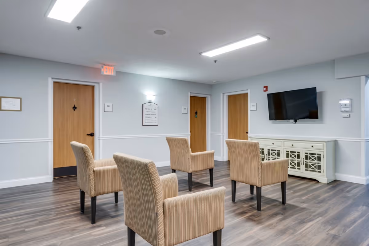 A common area with four beige upholstered armchairs arranged in a semi-circle on a wood floor. There are three wooden doors labeled 221, 222, and 223 on the light gray walls. A flat-screen TV is mounted on the wall above a white cabinet with glass-paneled doors. An exit sign is visible above one door, and a wall-mounted light fixture and a framed sign are also present.