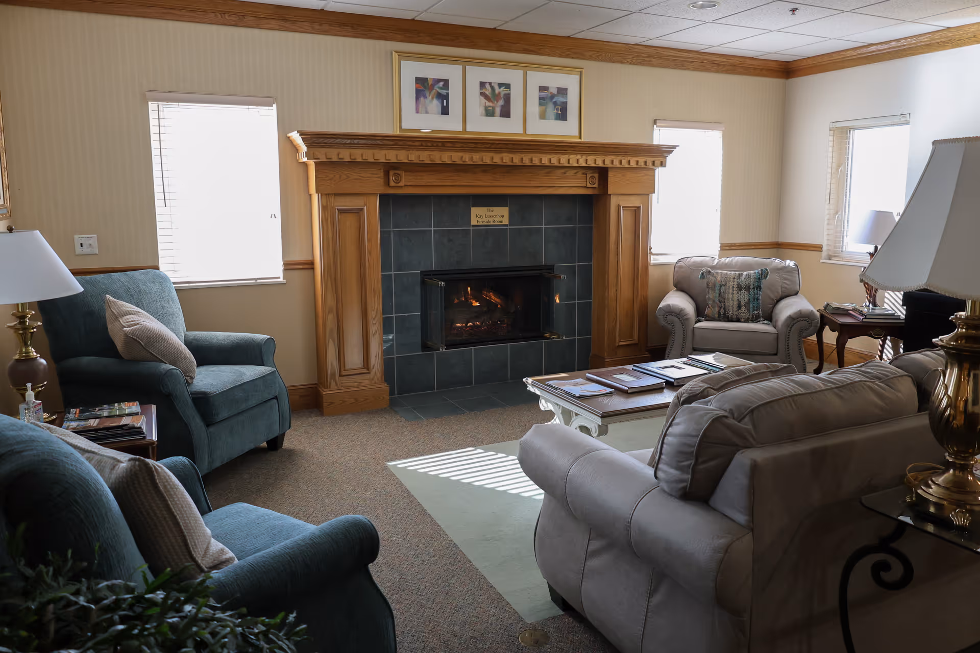 A cozy living room with a lit fireplace surrounded by a wooden mantle. The room has two blue armchairs, a beige armchair, and a beige sofa arranged around a white coffee table with books and magazines. There are three framed pictures above the fireplace and two windows with blinds letting in natural light. Lamps and side tables are placed beside the seating.