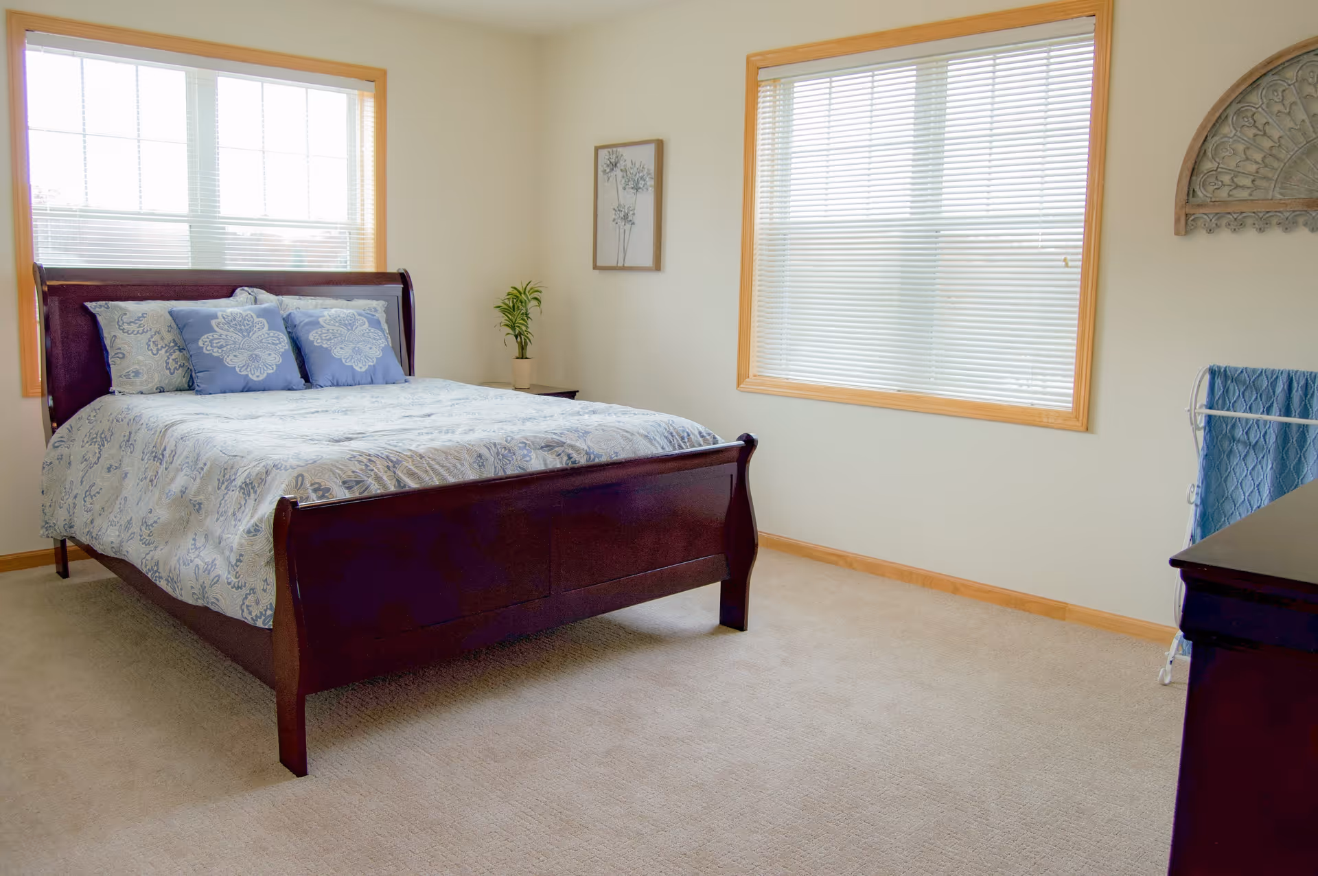 A bedroom with a wooden bed frame featuring a blue and white patterned bedspread and matching pillows. The room has beige carpet, two large windows with blinds, a small plant on a nightstand, a framed picture on the wall, and a towel rack with a blue towel.