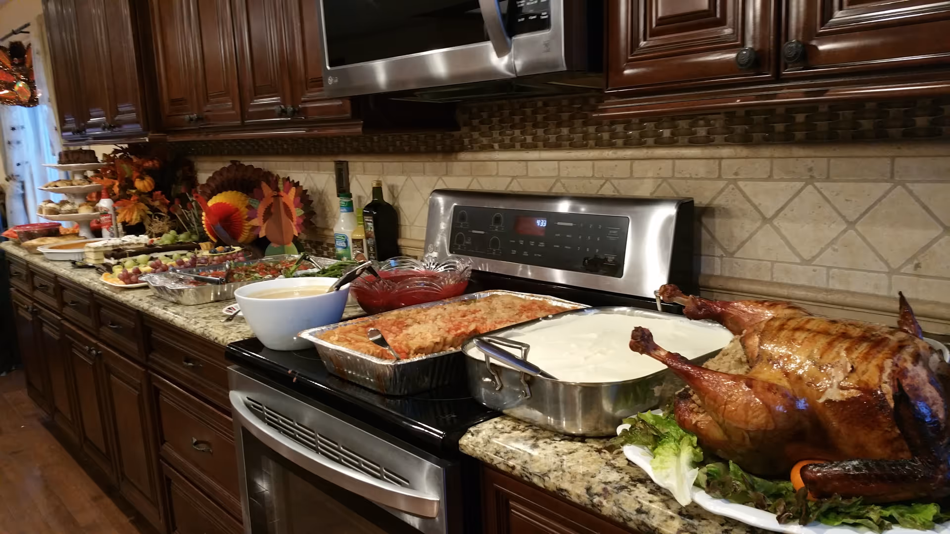 A kitchen countertop with a variety of food dishes including a roasted turkey, a large bowl of white sauce, a casserole dish, and other side dishes. The kitchen has dark wooden cabinets, a stainless steel stove and microwave, and a tiled backsplash. There are also decorative autumn-themed items on the counter.