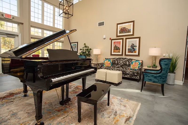Bright communal living room with a black grand piano, patterned sofa and chairs, artwork on the wall, and large windows.