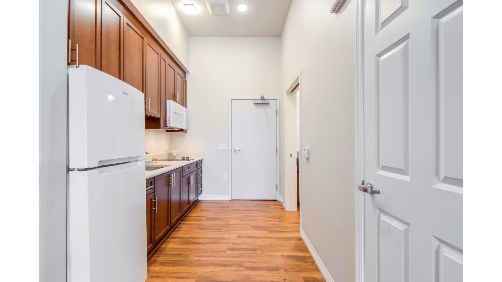 A narrow kitchen area with wooden cabinets and drawers on the left side, a white refrigerator, and a white microwave above the countertop. The floor is wooden, and there is a white door at the end of the kitchen space. Another white door is partially visible on the right side.