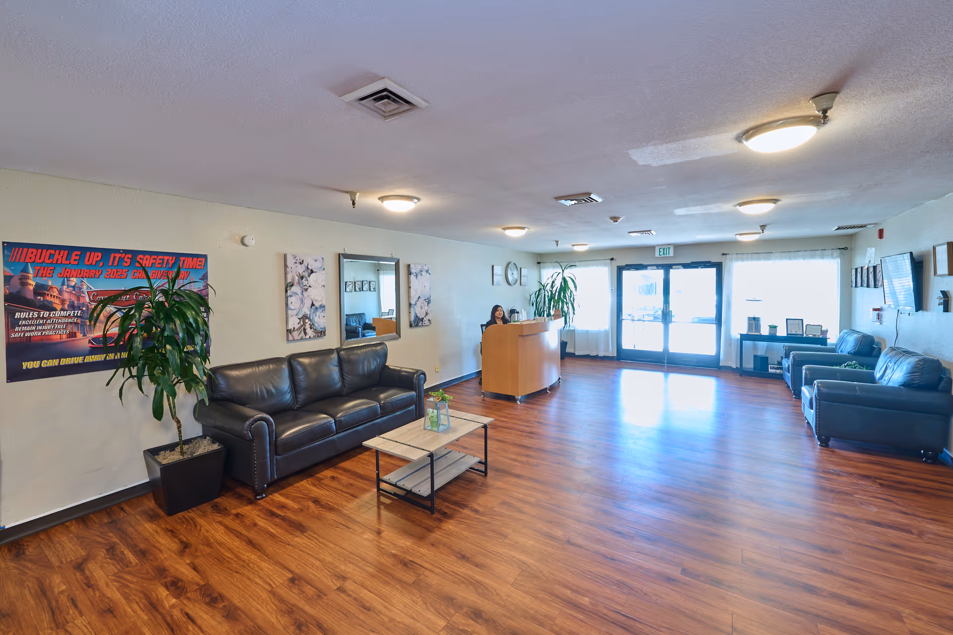 A spacious reception area with wooden flooring, featuring a woman standing behind a curved wooden reception desk. The room has several black leather sofas, a coffee table with a small plant, a large potted plant, wall decorations including mirrors and a safety poster, and large windows with sheer curtains letting in natural light.