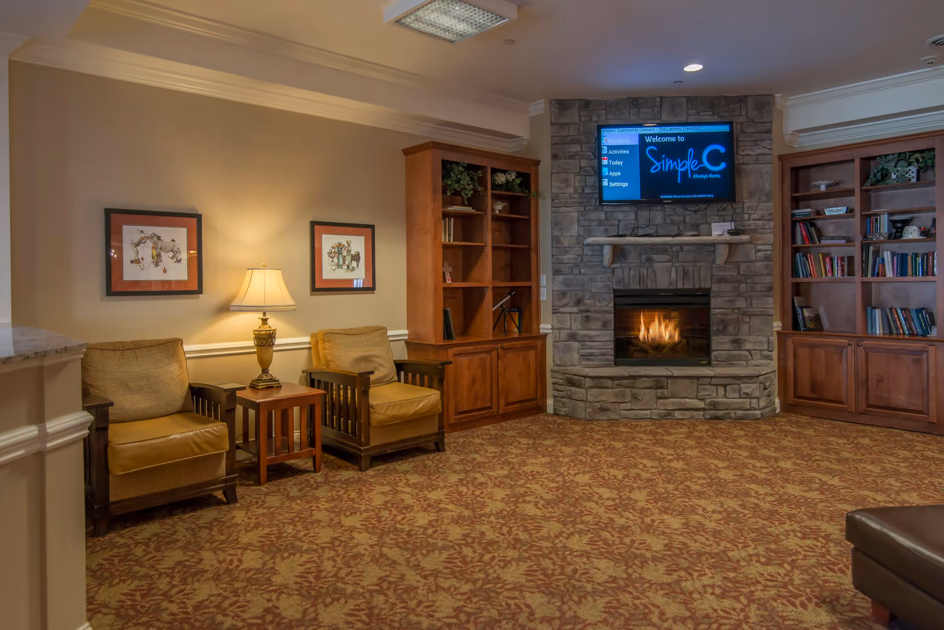 A cozy living room area with two cushioned armchairs separated by a wooden side table with a lamp. The walls are decorated with two framed pictures. There are two wooden bookshelves flanking a stone fireplace with a fire burning inside. Above the fireplace is a mounted flat-screen TV displaying a welcome message for SimpleC. The room has patterned carpet flooring and soft lighting.