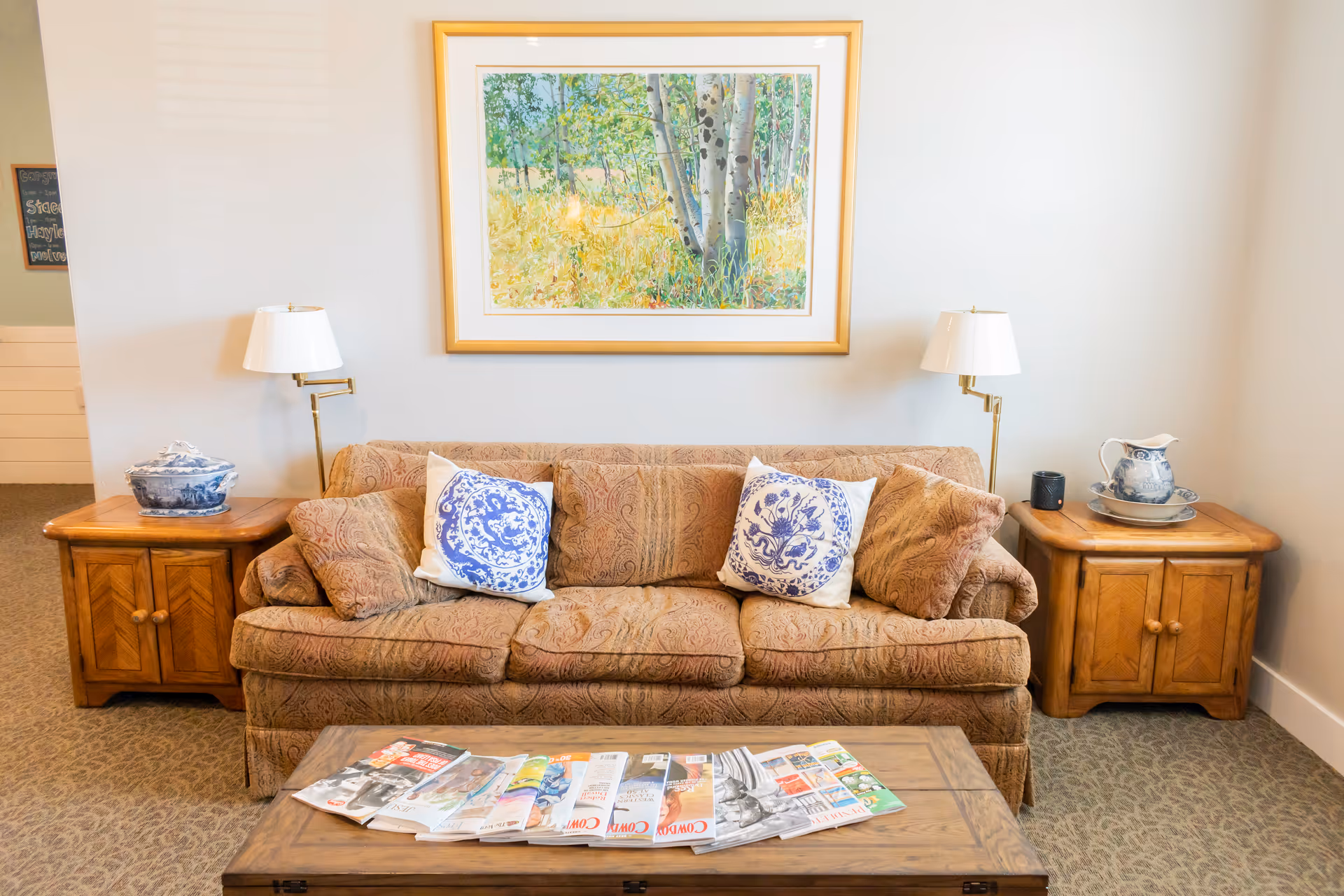 A cozy living room area with a patterned brown sofa adorned with two white and blue decorative pillows and two matching brown pillows. On either side of the sofa are wooden side tables, each with a white lamp and blue and white ceramic decor pieces. Above the sofa hangs a framed painting of a forest scene with trees and greenery. In front of the sofa is a wooden coffee table with various magazines spread out on top. The room has light-colored walls and carpeted flooring.