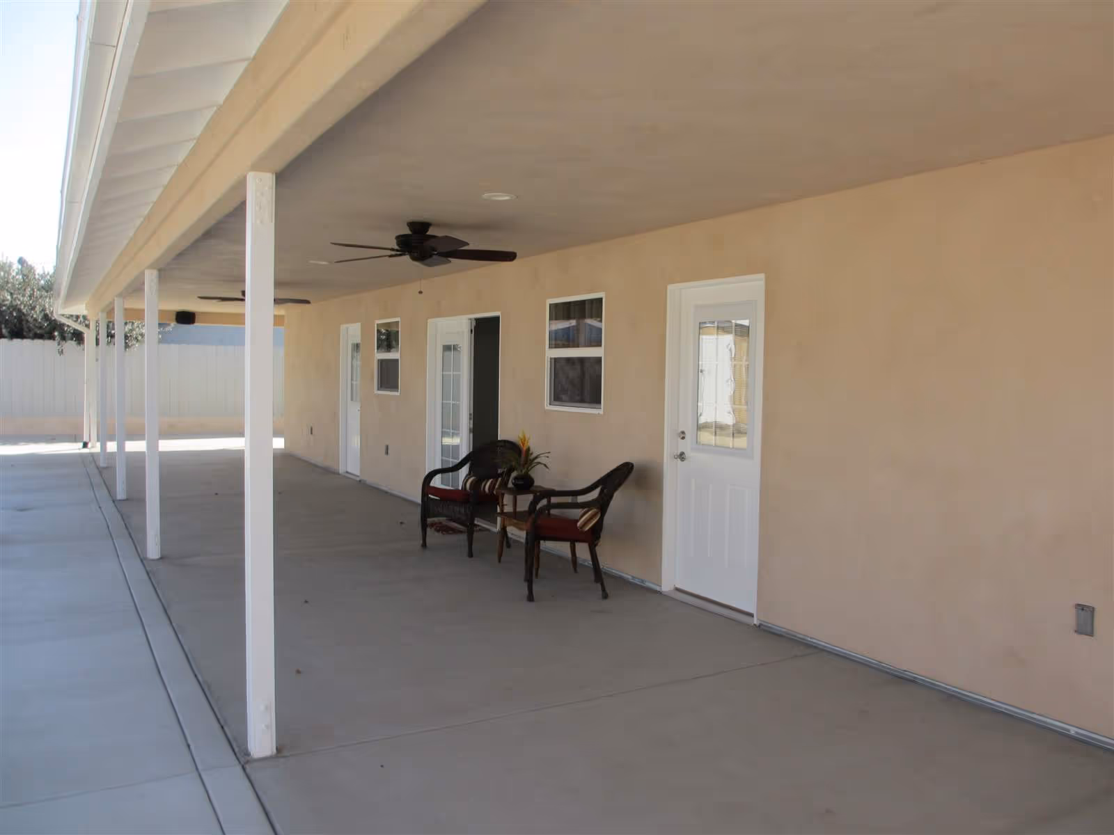 Covered outdoor patio area with beige walls, white doors, two wicker chairs with red cushions, a small table with a plant, ceiling fans, and white support columns.