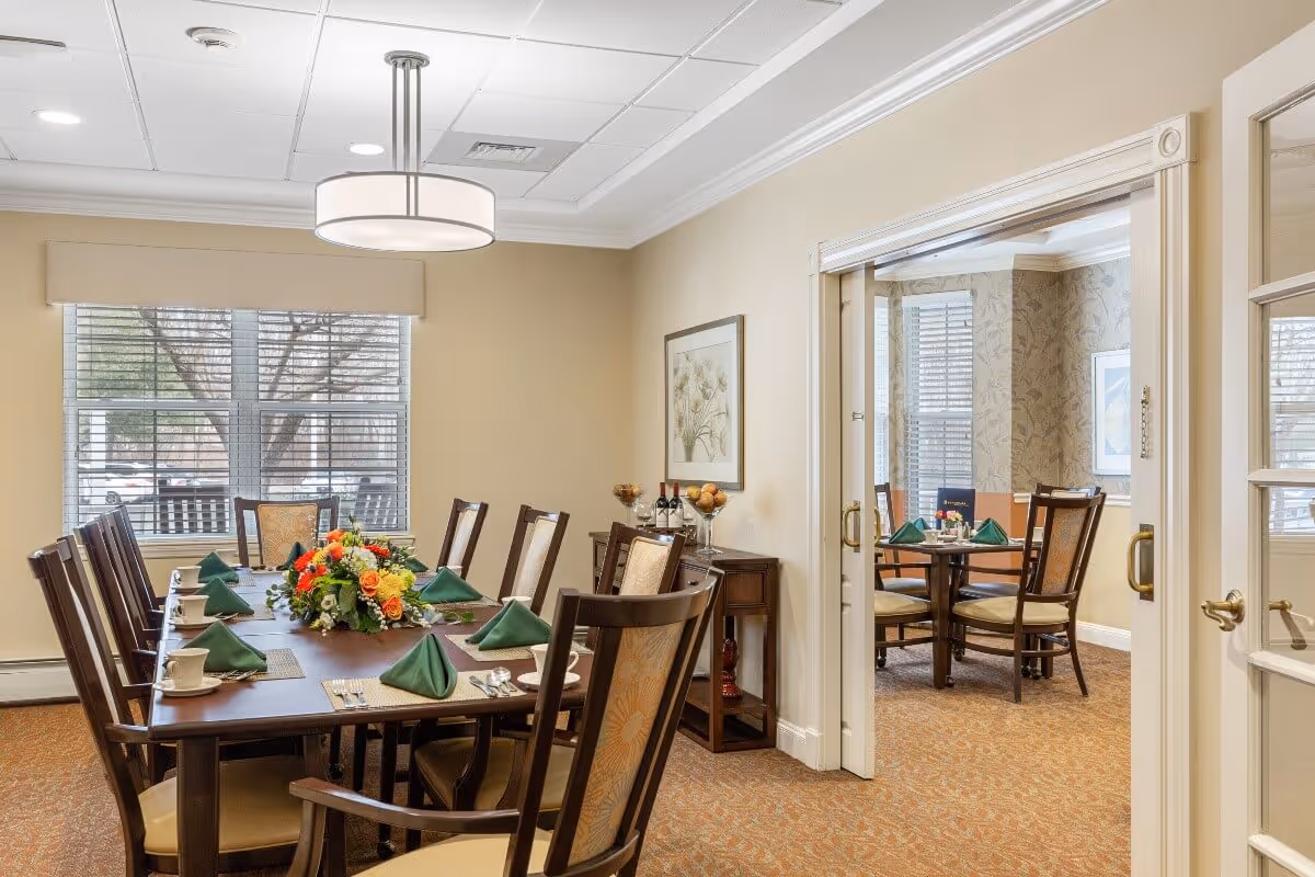 A well-lit dining room with a large wooden table set for eight people, featuring green folded napkins, white cups, and a floral centerpiece. The room has beige walls, a large window with blinds, and a modern ceiling light fixture. Through an open doorway, another smaller dining area with a round table and four chairs is visible, also set with green napkins and tableware.