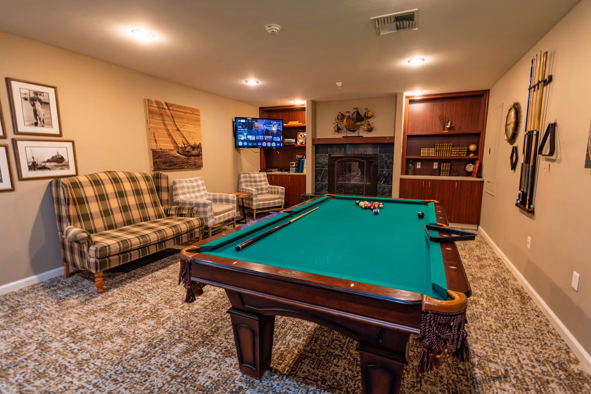 Game room with a green pool table, plaid sofa and armchairs, a wall-mounted TV, fireplace and built-in shelving.