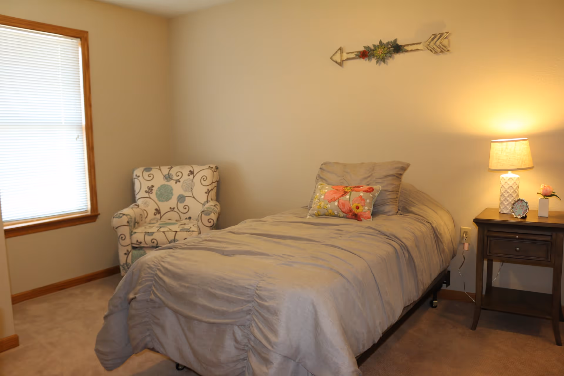 A cozy bedroom with a single bed covered in a gray comforter and a floral pillow. Next to the bed is a wooden nightstand with a lamp, a small flower vase, and a decorative item. A patterned armchair is placed near a window with closed blinds. A decorative arrow hangs on the wall above the bed.