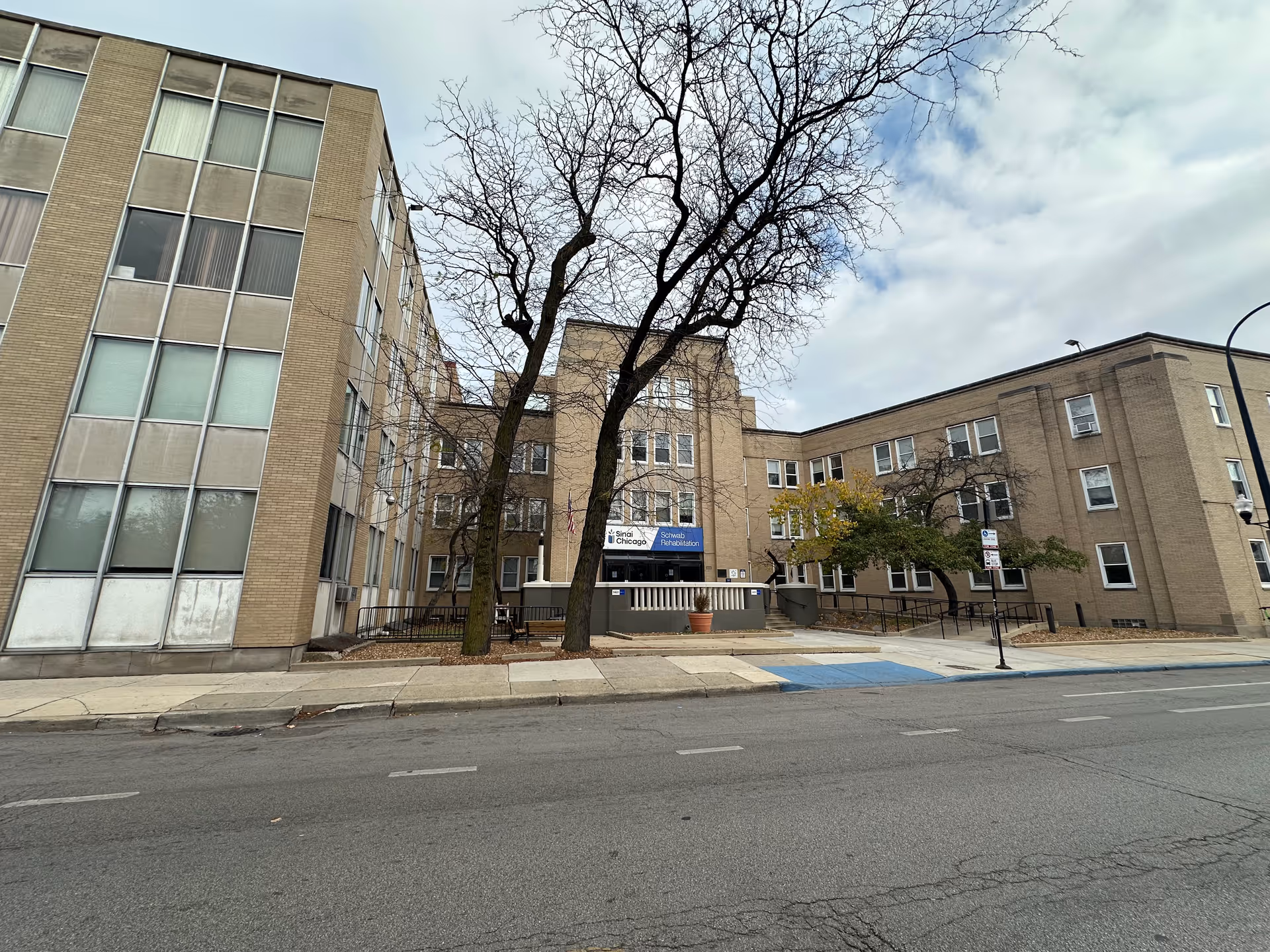 Front view of a multi-story brick rehabilitation center with leafless trees and an entrance ramp under a cloudy sky.